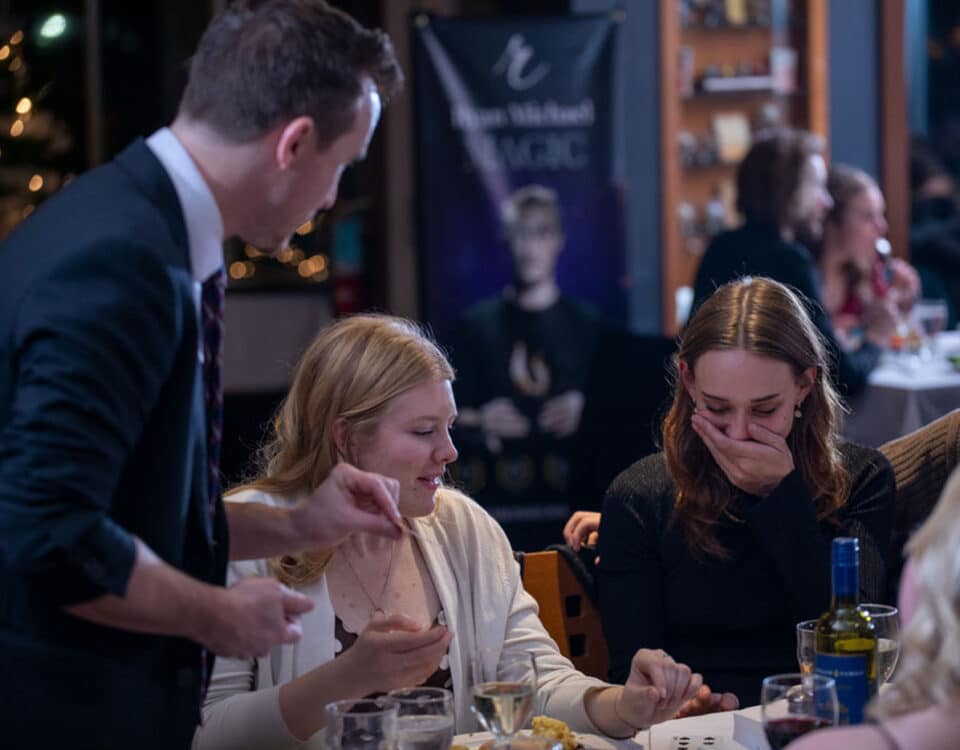A magician performs at a dining table, with two women reacting with surprise and laughter while a man assists them.
