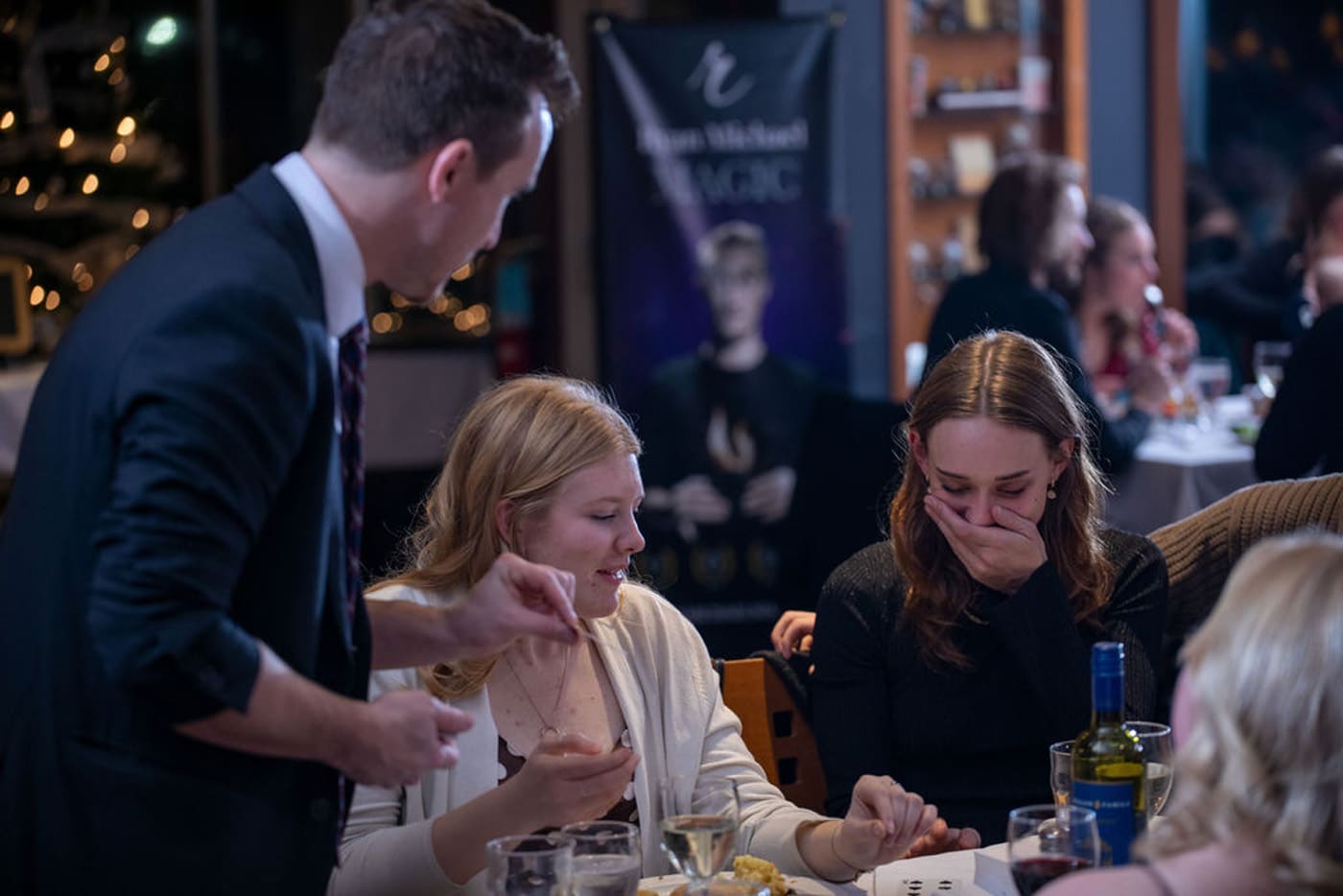 A magician performs at a dining table, with two women reacting with surprise and laughter while a man assists them.
