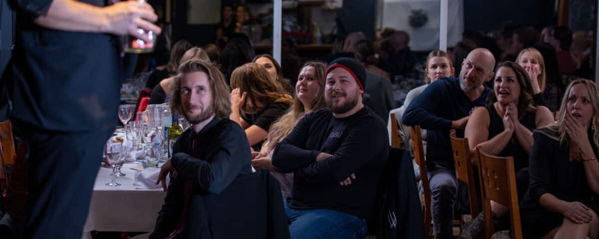A magician performing for an audience at an event, with guests reacting with laughter and surprise.