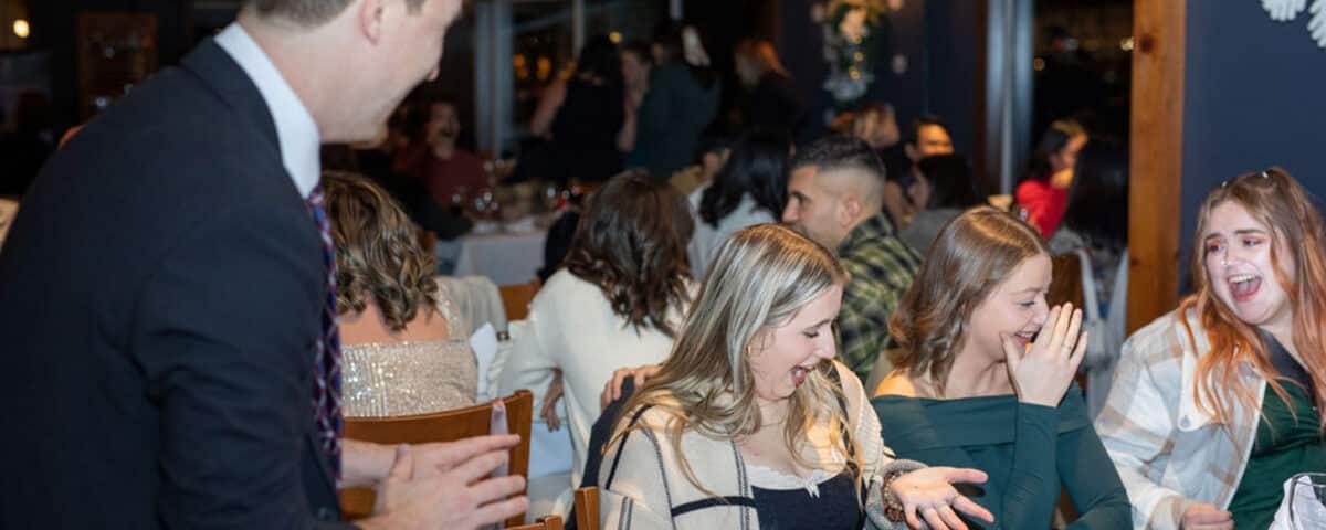 A group of people is sitting at a dining table, enjoying themselves during a celebration. A man in a suit is engaging with a few women at the table, who are smiling and laughing.
