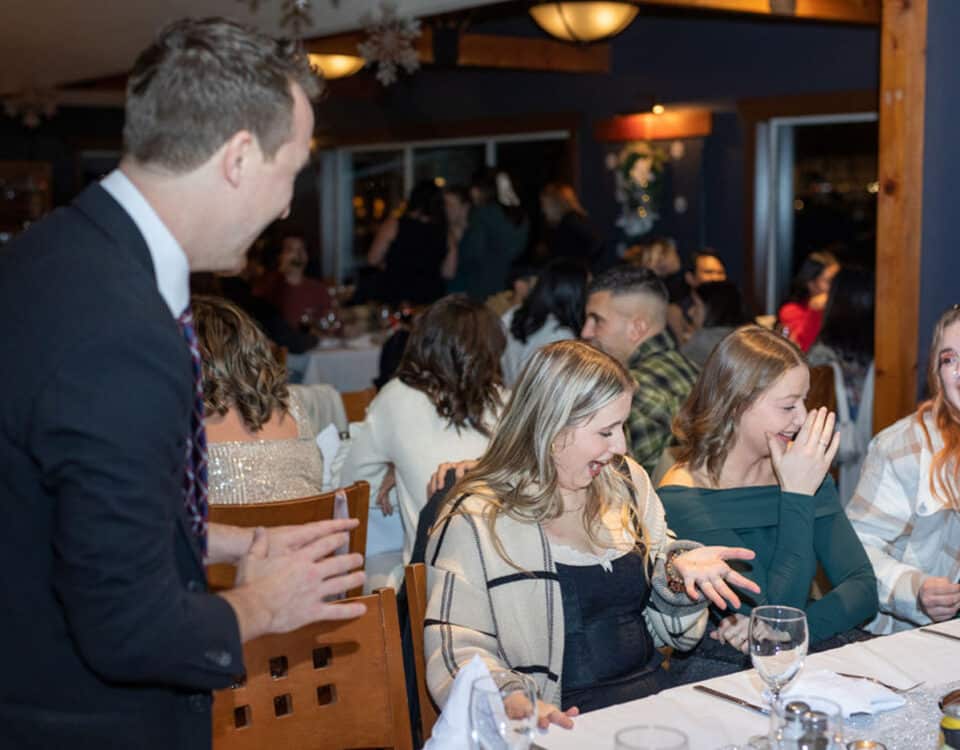 A group of people is sitting at a dining table, enjoying themselves during a celebration. A man in a suit is engaging with a few women at the table, who are smiling and laughing.