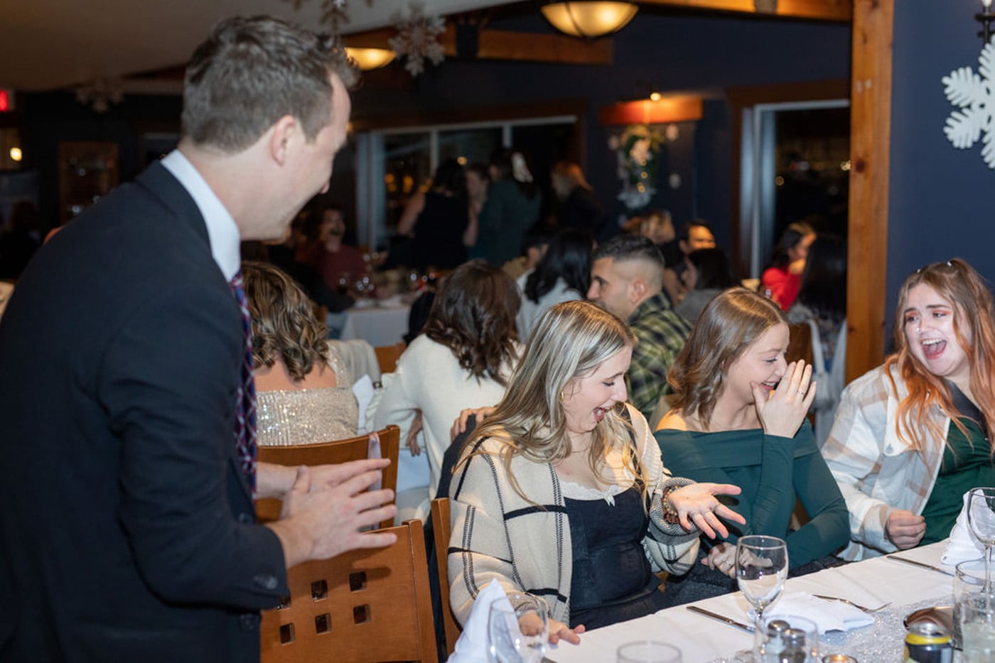 A group of people is sitting at a dining table, enjoying themselves during a celebration. A man in a suit is engaging with a few women at the table, who are smiling and laughing.