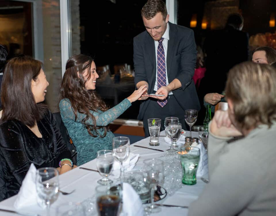 A magician performs a card trick for a smiling woman at a dining table, while other guests watch. The table is set with drinks and place settings.