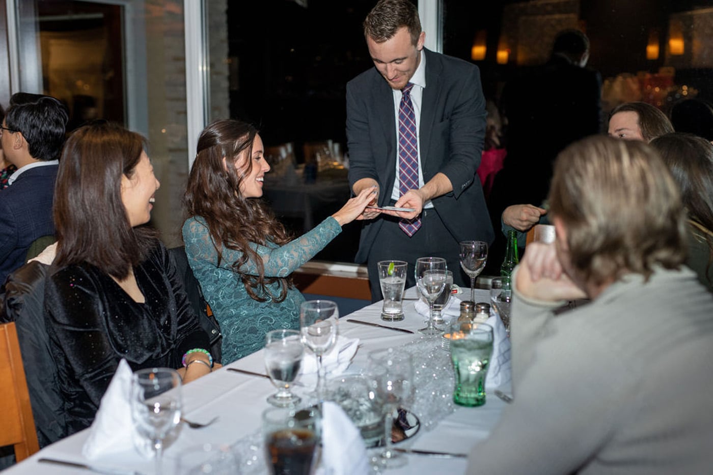 A magician performs a card trick for a smiling woman at a dining table, while other guests watch. The table is set with drinks and place settings.
