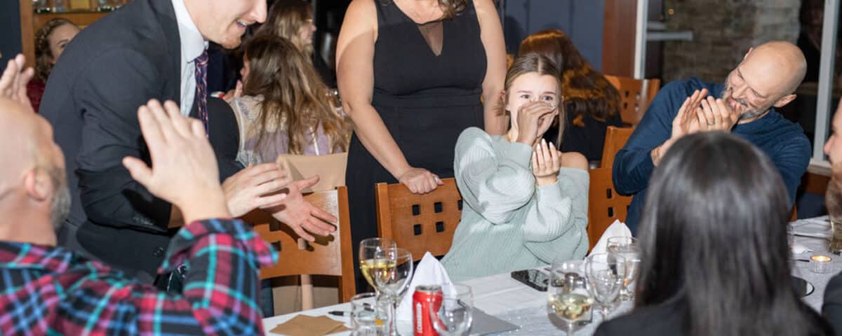 A group of people seated at a dinner table, with a man in a suit entertaining them. A woman in a black dress is smiling while another woman, dressed in a sweater, appears amused. Drinks and plates are on the table.