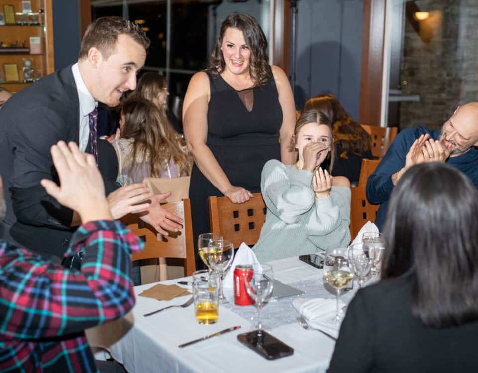A group of people seated at a dinner table, with a man in a suit entertaining them. A woman in a black dress is smiling while another woman, dressed in a sweater, appears amused. Drinks and plates are on the table.