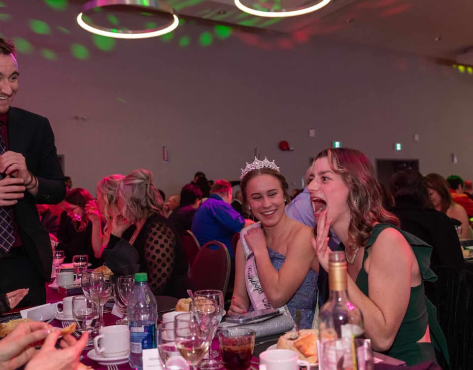 A lively dinner scene with several people at a table, including two women laughing, one wearing a tiara and an evening gown.
