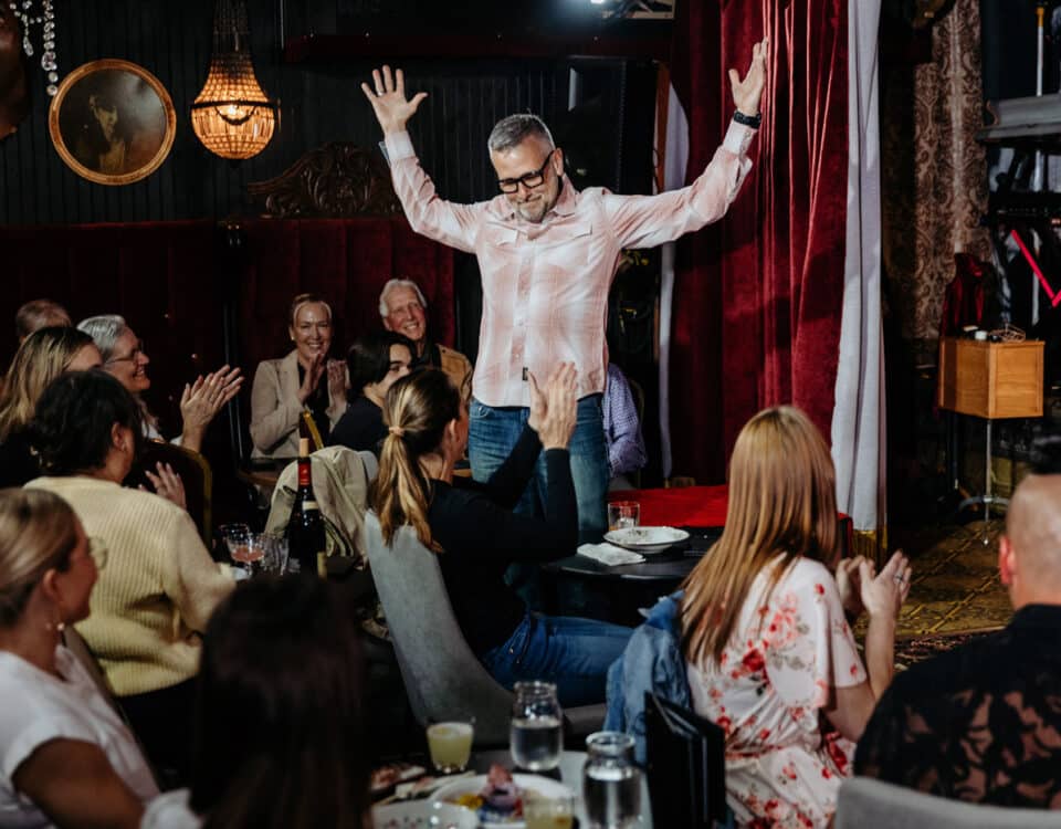 A man with glasses and a plaid shirt is joyfully raising his arms on stage, while a seated audience applauds, showing expressions of enjoyment in a lively venue.