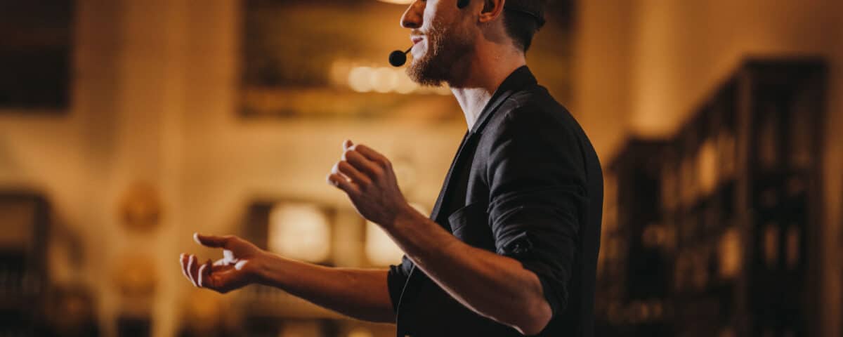 A man speaking passionately with a microphone headset, gesturing with his hands in a warmly lit setting.