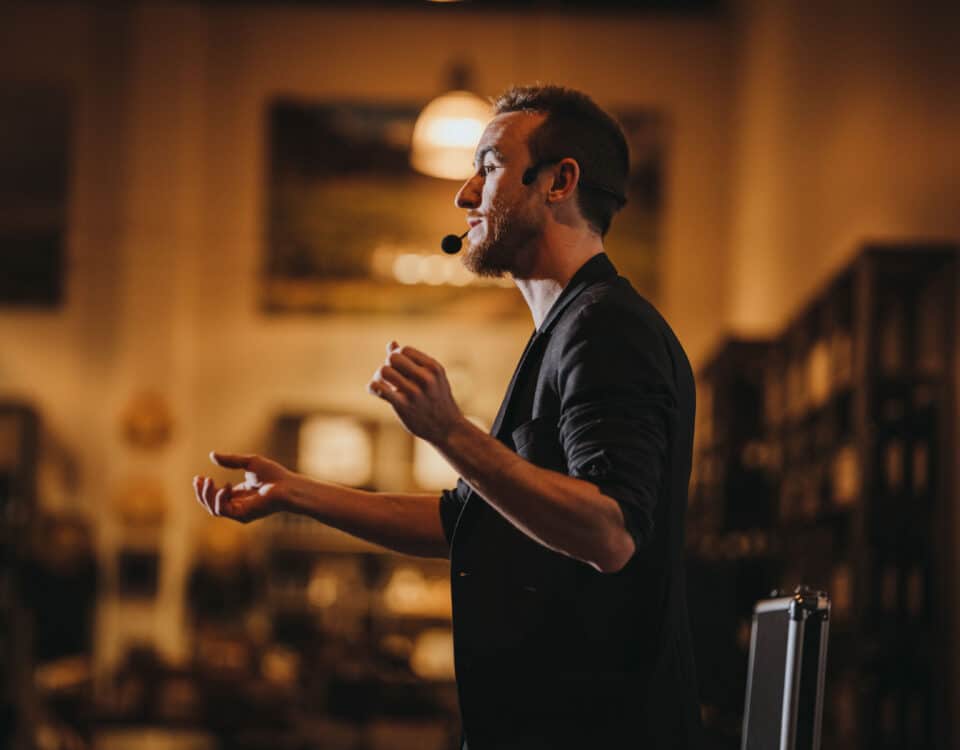 A man speaking passionately with a microphone headset, gesturing with his hands in a warmly lit setting.