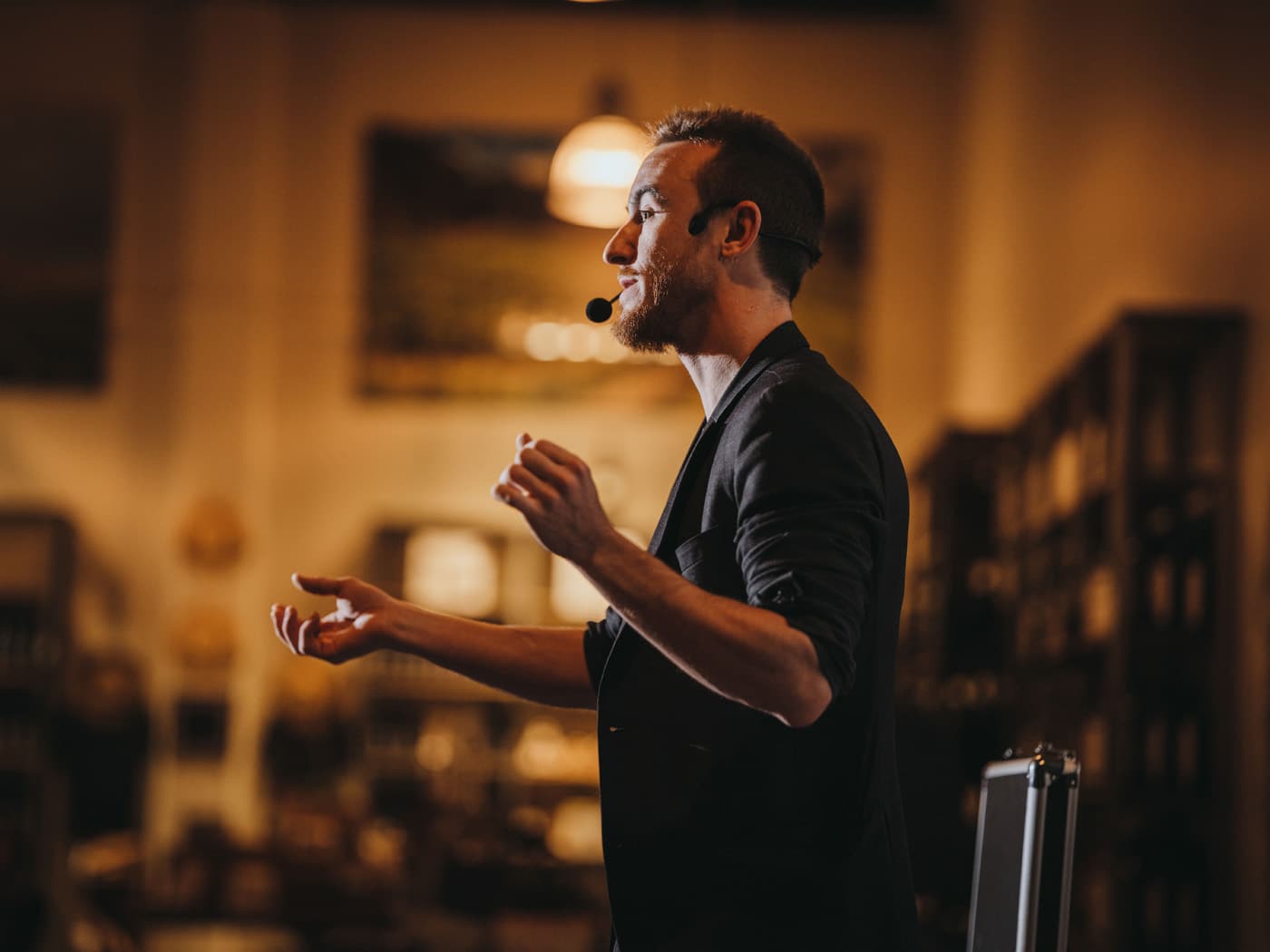 A man speaking passionately with a microphone headset, gesturing with his hands in a warmly lit setting.