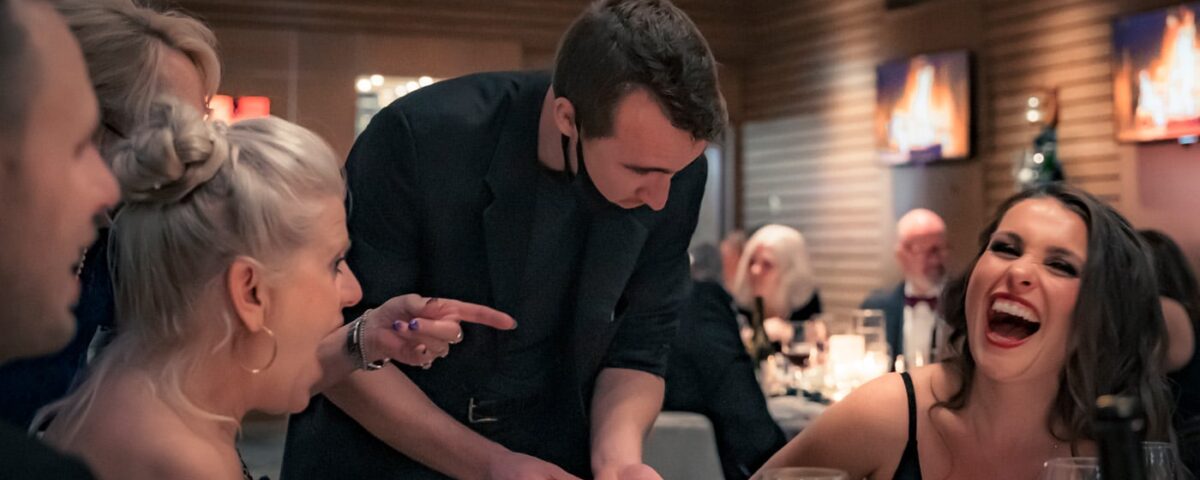 A magician performs a trick at a lively dinner, entertaining a group of mostly smiling guests, including two women who are reacting with surprise and laughter. Glasses of drinks are on the table.
