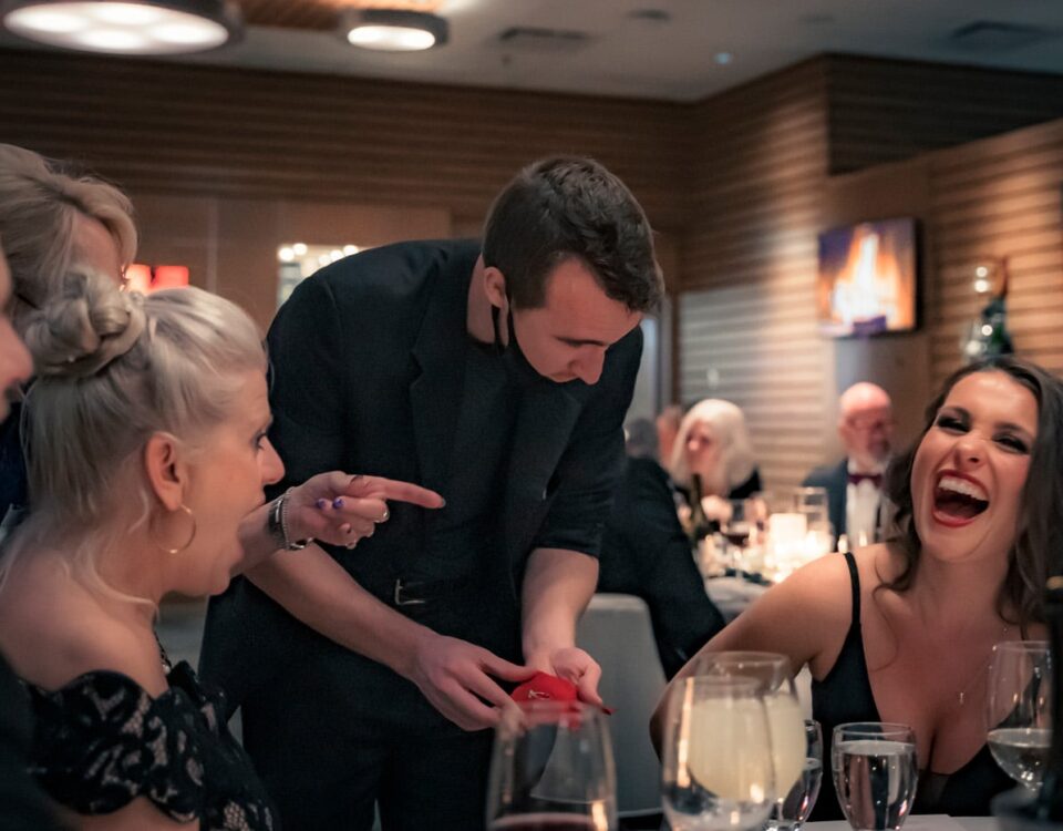 A magician performs a trick at a lively dinner, entertaining a group of mostly smiling guests, including two women who are reacting with surprise and laughter. Glasses of drinks are on the table.