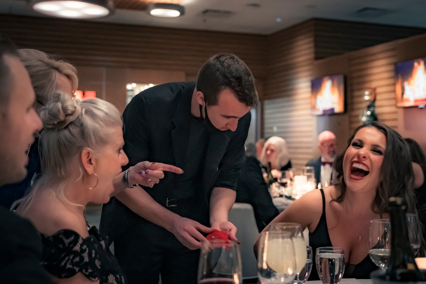 A magician performs a trick at a lively dinner, entertaining a group of mostly smiling guests, including two women who are reacting with surprise and laughter. Glasses of drinks are on the table.