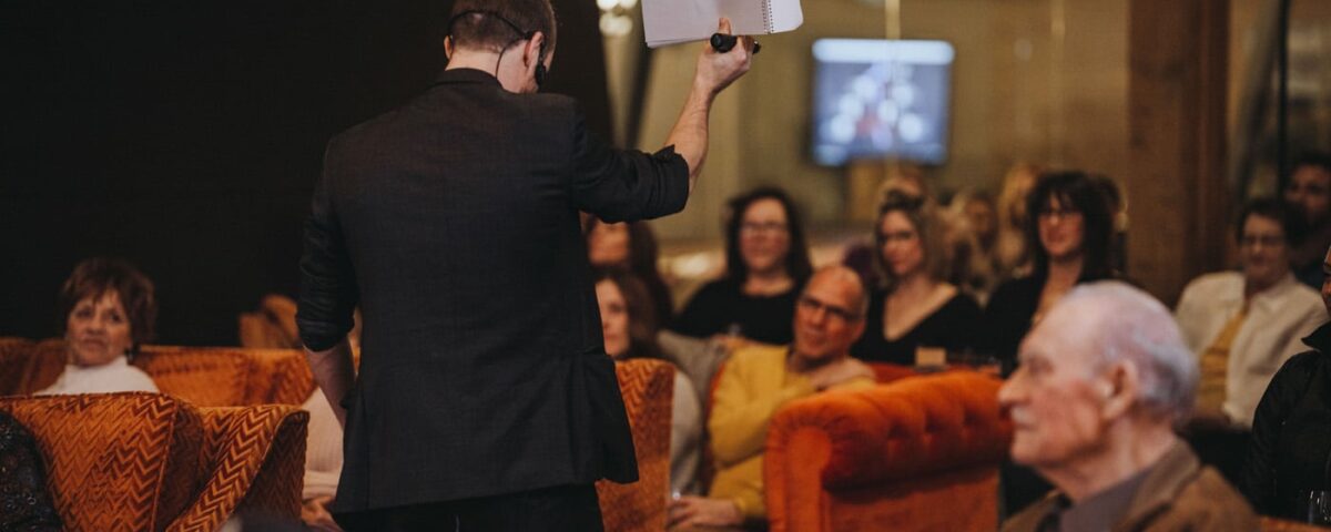 A speaker holds up a paper while addressing an audience seated in orange couches, with several attendees focused on the presentation.