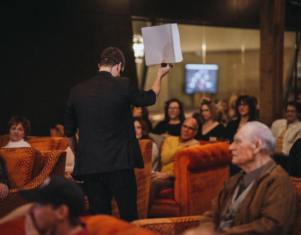 A speaker holds up a paper while addressing an audience seated in orange couches, with several attendees focused on the presentation.