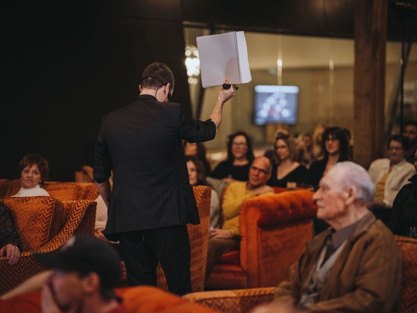 A speaker holds up a paper while addressing an audience seated in orange couches, with several attendees focused on the presentation.