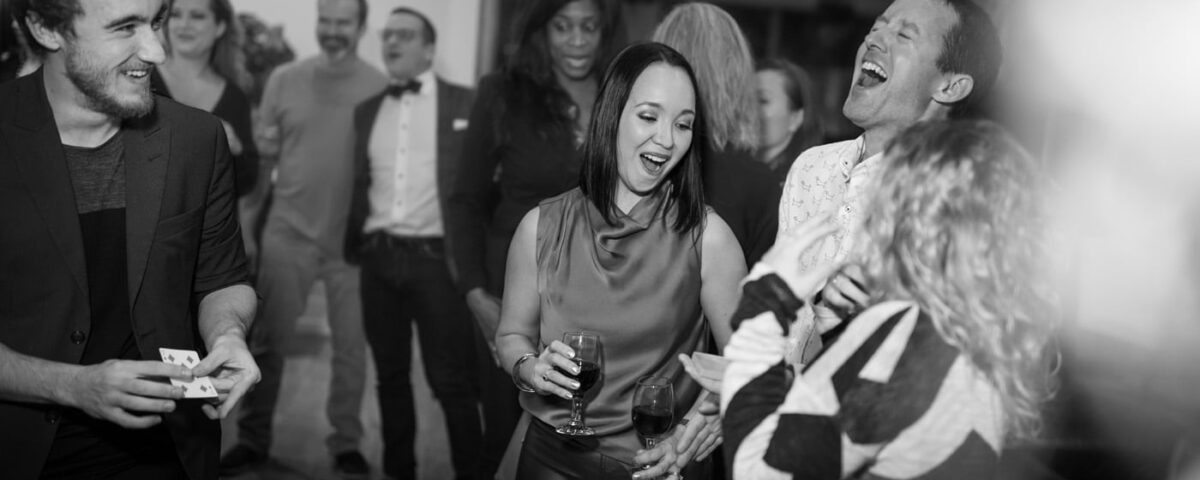 A lively black-and-white image of a group of people enjoying a social gathering, with some laughing and interacting, while one person performs magic tricks.