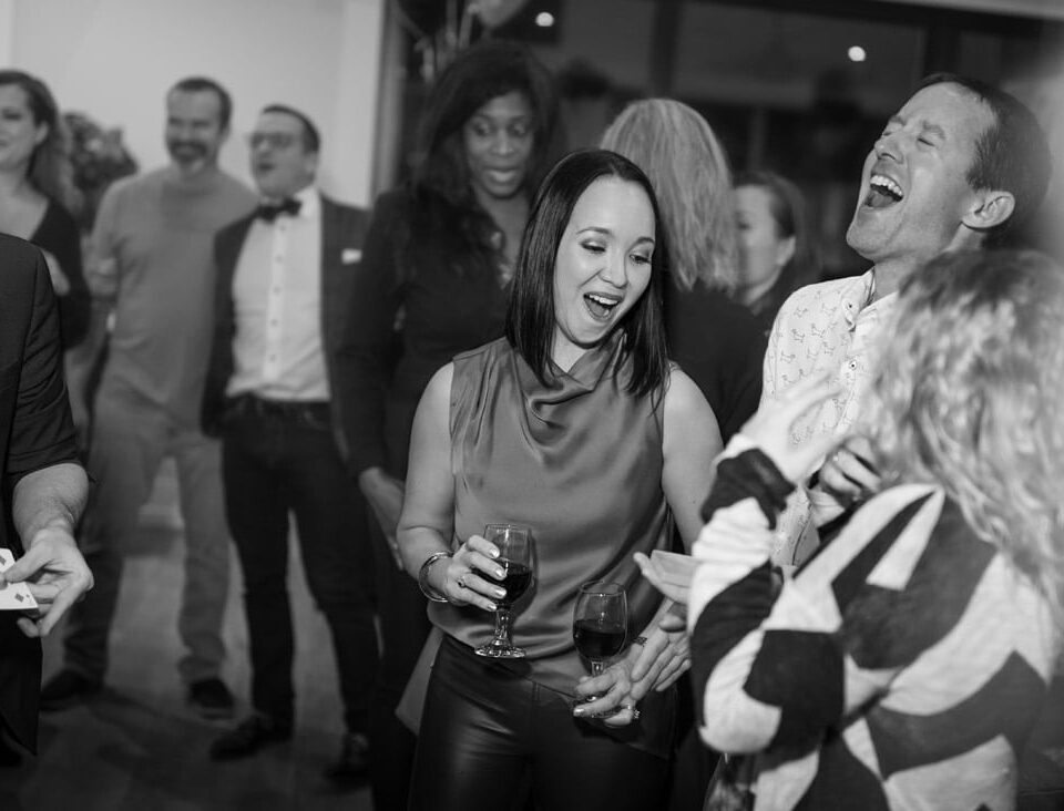 A lively black-and-white image of a group of people enjoying a social gathering, with some laughing and interacting, while one person performs magic tricks.