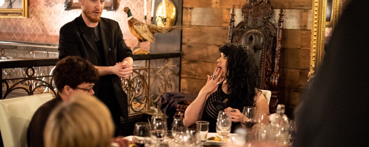 A group of people at a dining table, with one person gesturing while speaking and another looking surprised. The setting features elegant decor and tableware.