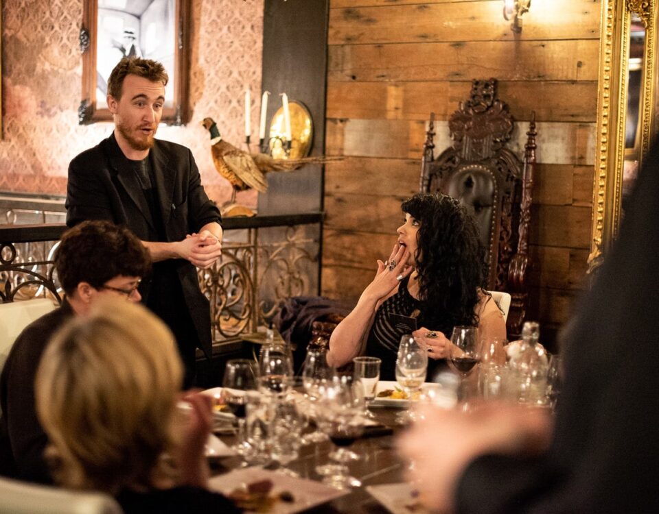A group of people at a dining table, with one person gesturing while speaking and another looking surprised. The setting features elegant decor and tableware.