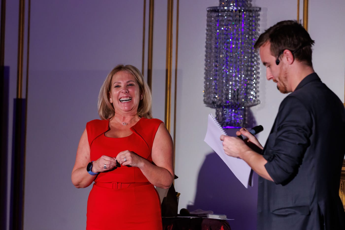 A woman in a red dress smiles and laughs while standing on stage, as a man in a black jacket takes notes. A chandelier is visible in the background.