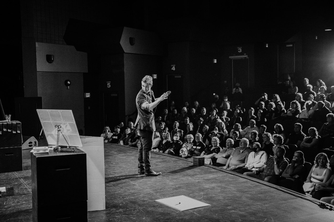 A performer standing on stage in front of a seated audience, gesturing as they speak. The scene is in black and white, with a presentation board nearby.