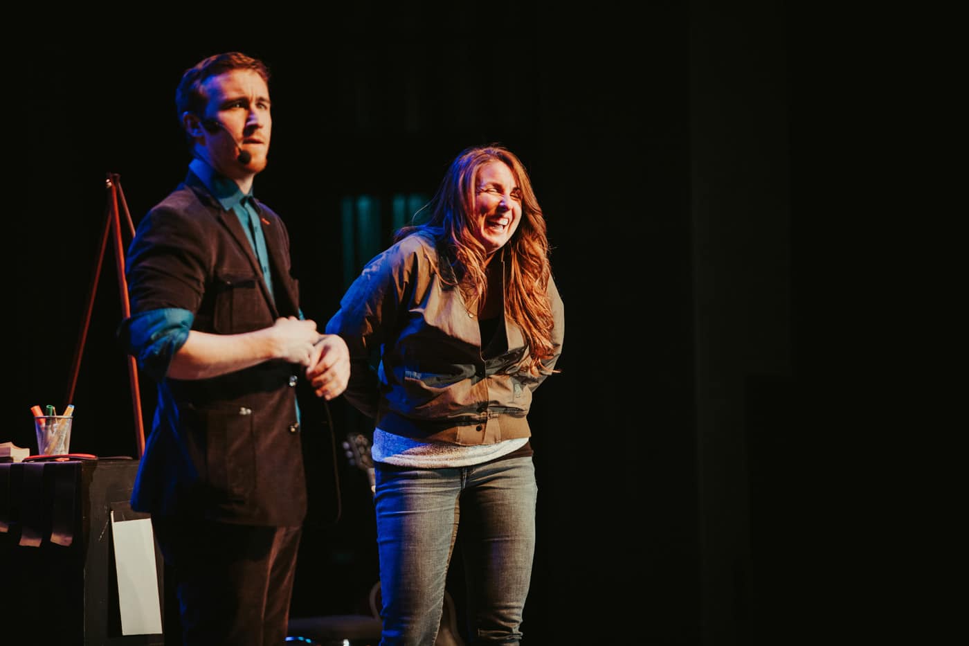 A man and a woman are performing on stage. The woman is laughing, while the man appears to be speaking or engaging the audience. There are props on a table beside them.