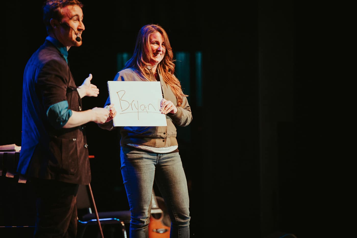 A performer stands on stage with a microphone, holding a sign that reads "Bryan." A woman next to him is smiling and holding the sign.