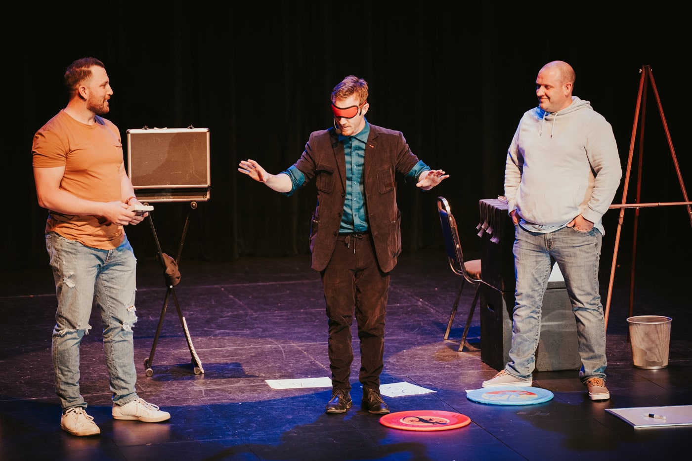 Three men perform on stage, with one man in the center wearing a blindfold and gesturing, while the others observe.