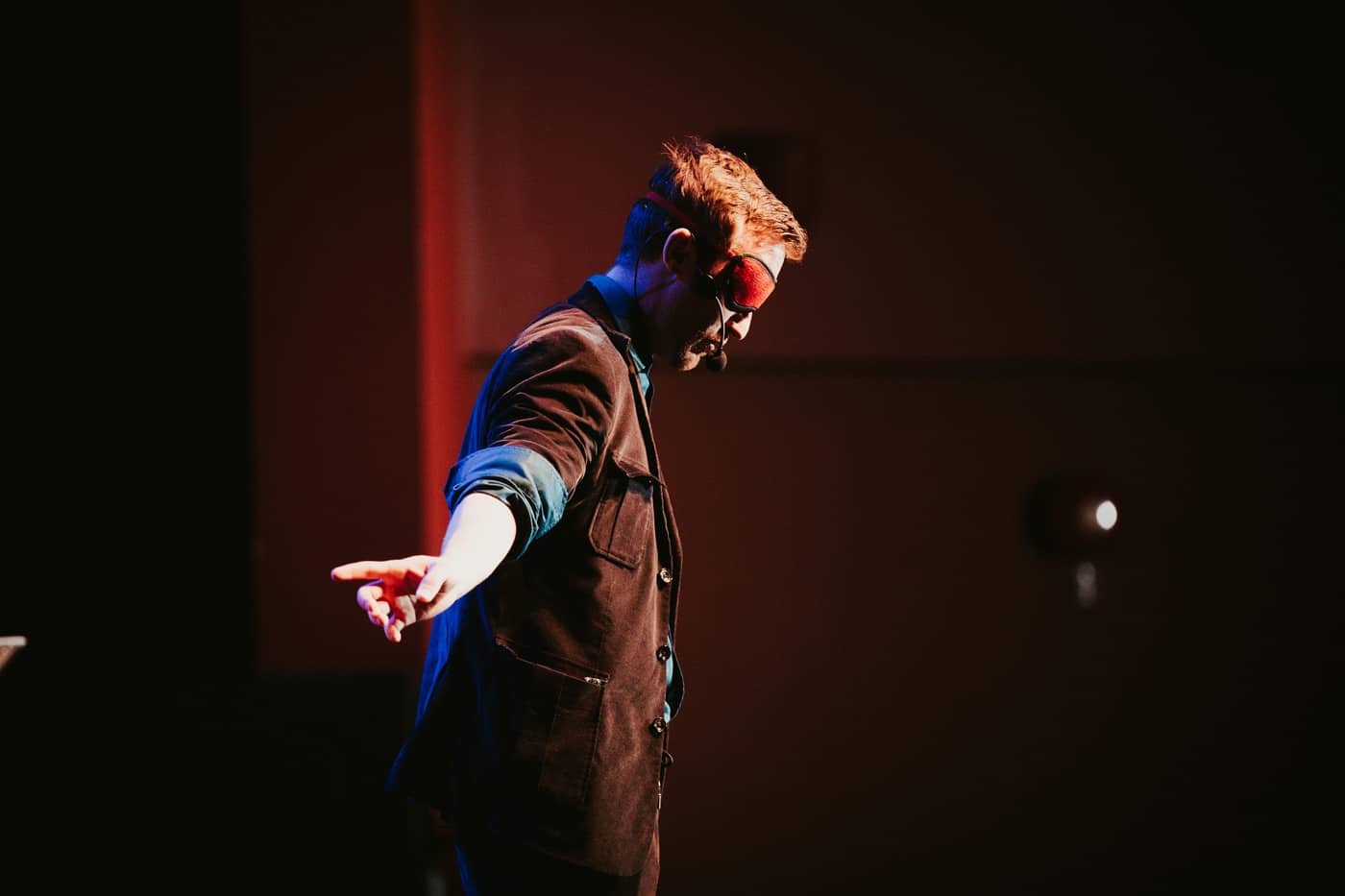A performer wearing a blindfold stands with one arm extended, against a dimly lit background.
