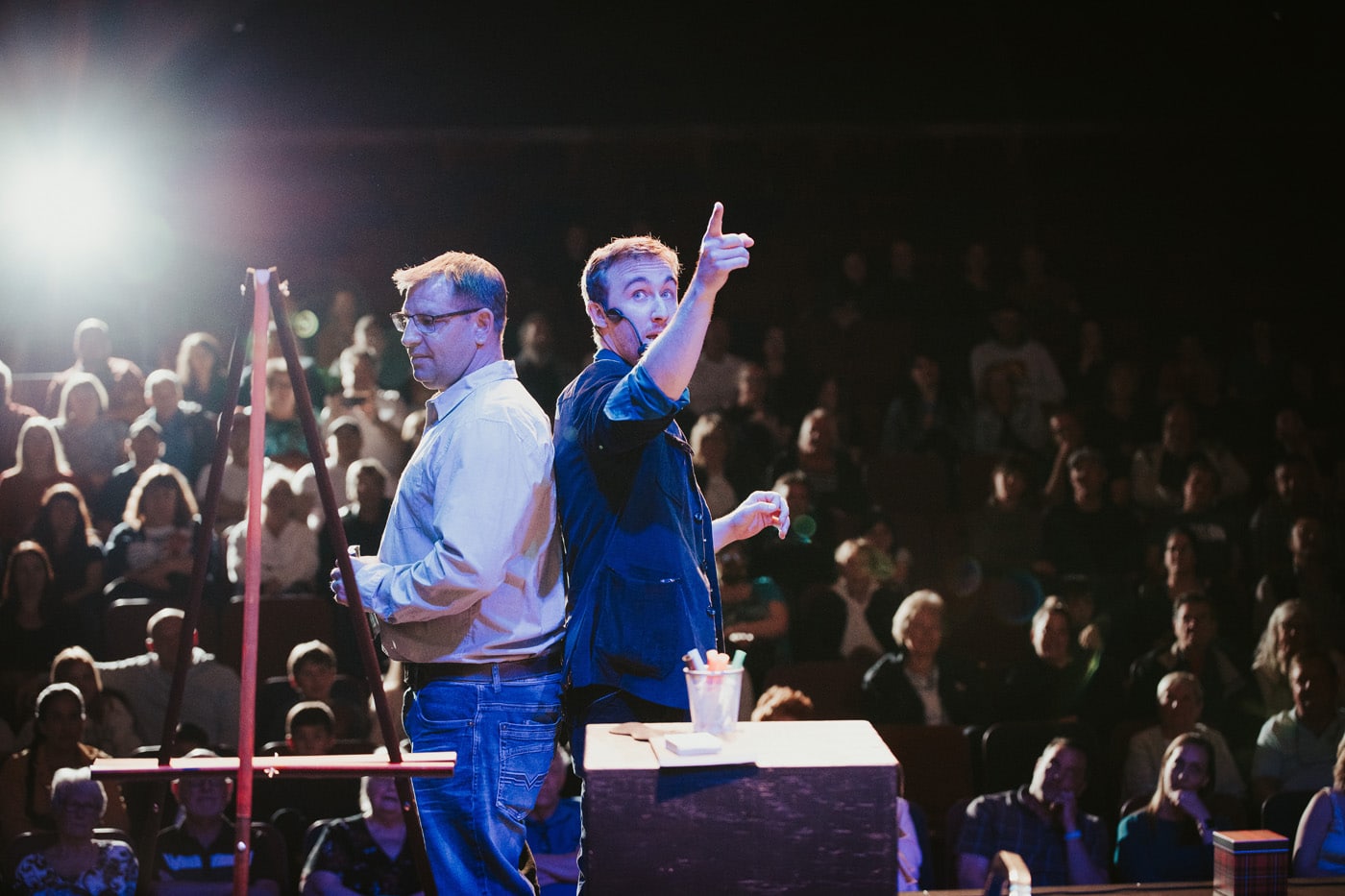 Two men on stage, one pointing and speaking to the audience, while the other stands still.