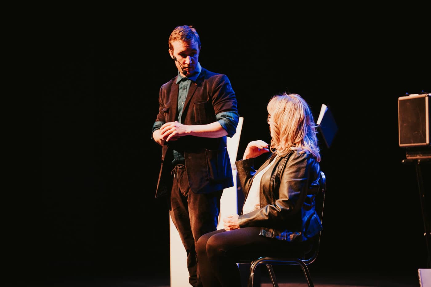 A performer interacts with a seated woman on stage, with a dark background and spotlighting.