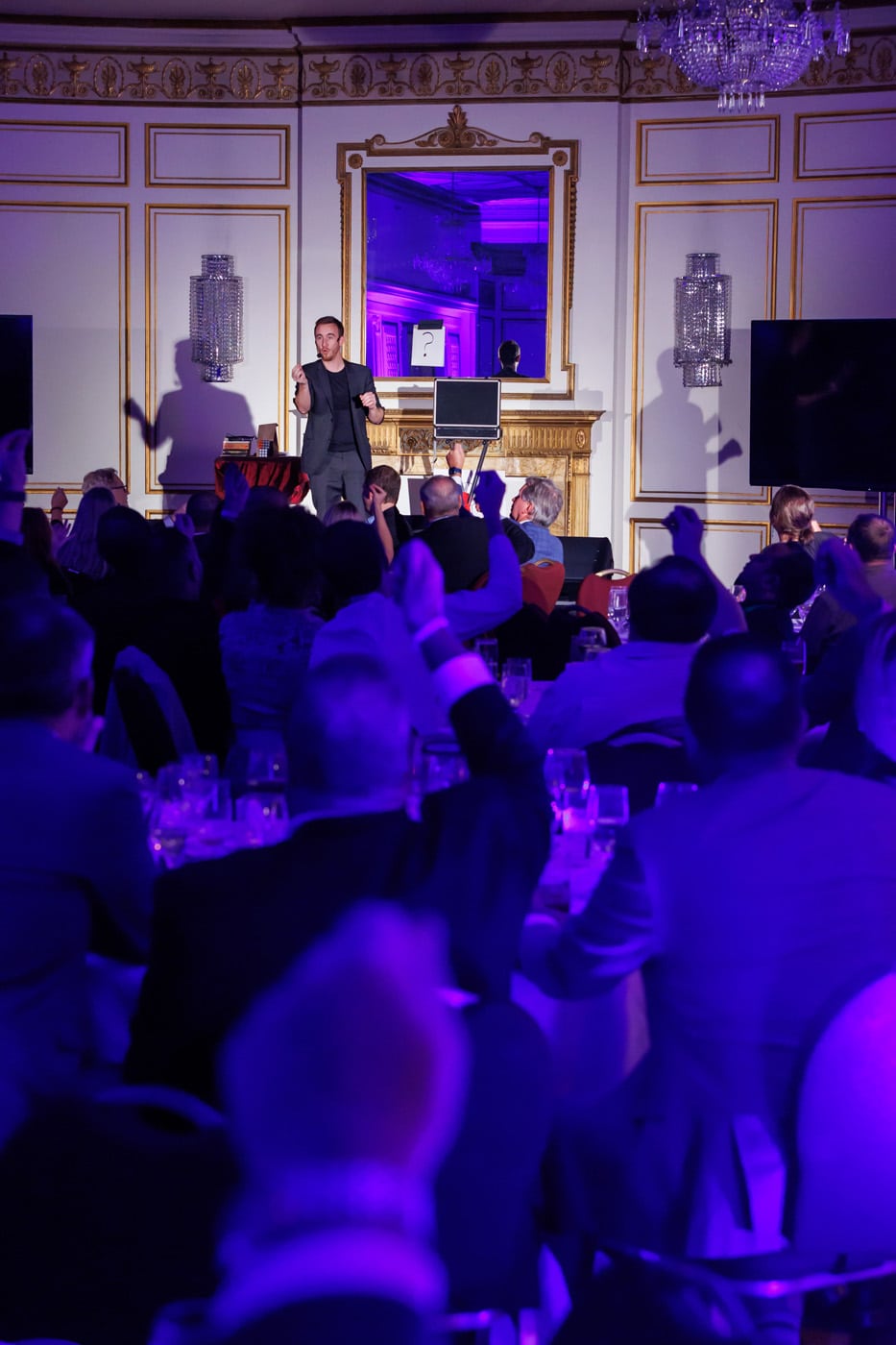 A performer on stage engages with an audience in a well-decorated room, with hands raised and a spotlight illuminating the scene.