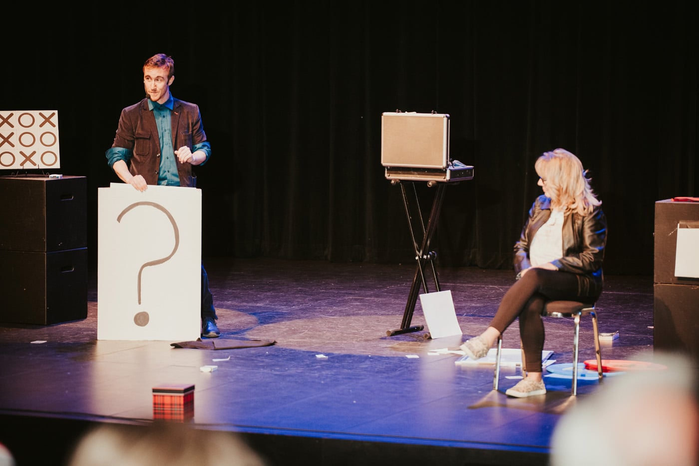 A magician performs on stage, holding a large sign with a question mark, while a woman sits nearby on a chair. The scene includes a tic-tac-toe board and scattered papers on the floor.