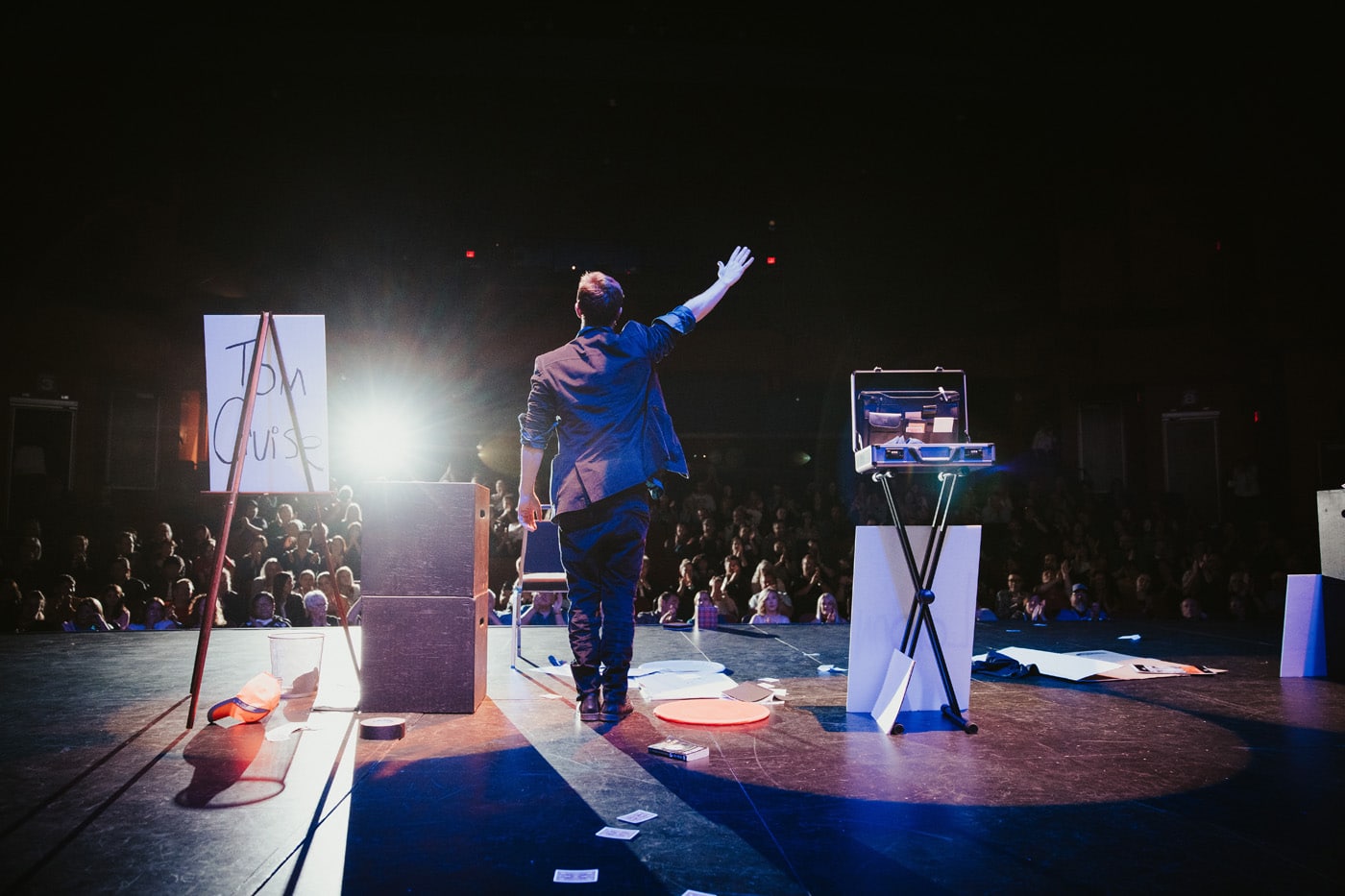 A performer on stage gestures towards an audience, with a sign reading "Tom Cruise" nearby and various props scattered on the floor.