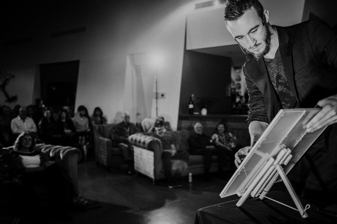 A performer stands in front of an audience, focused on an easel while engaging with the crowd in a dimly lit venue.