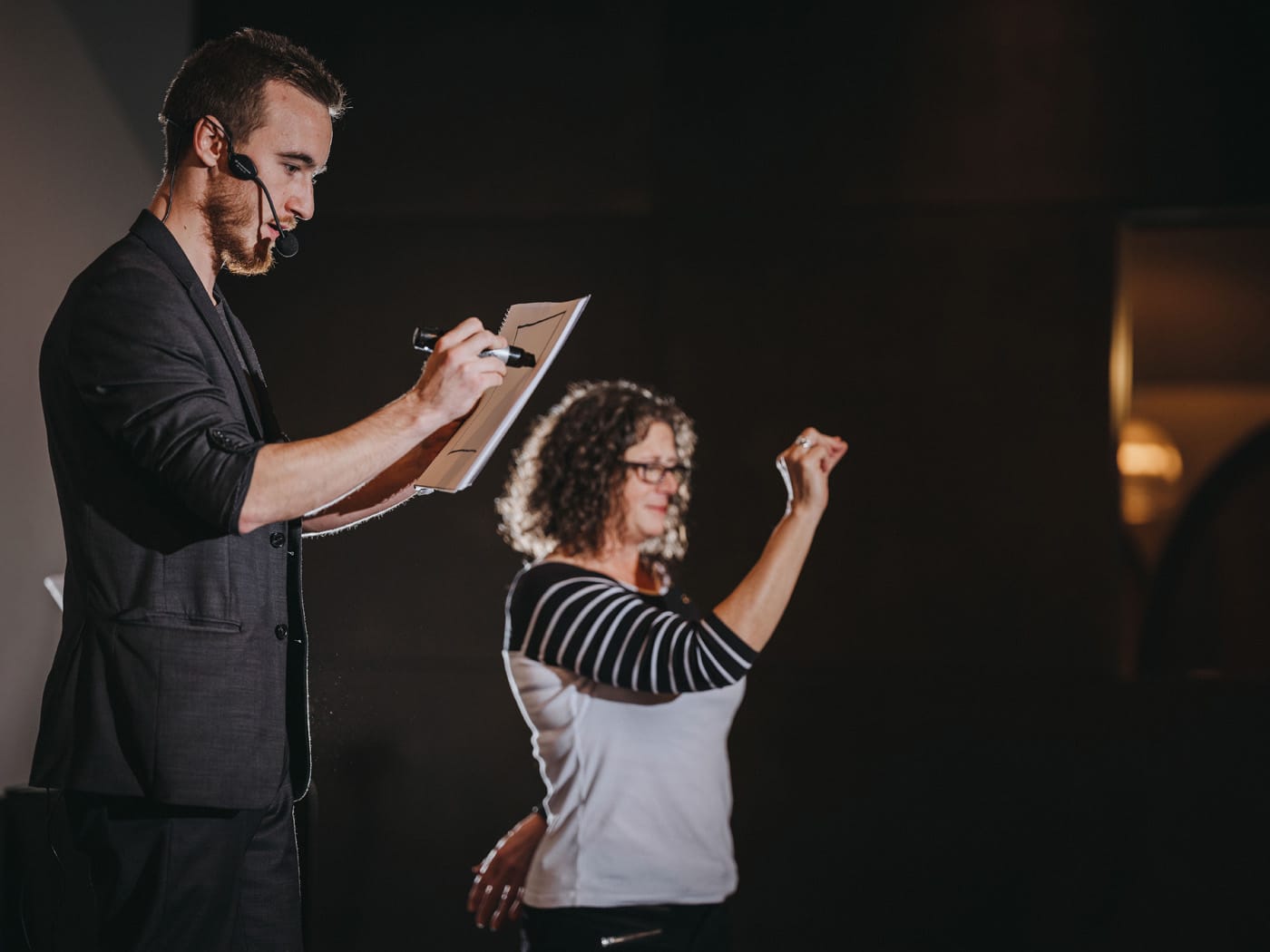 A man in a suit is drawing on a pad with a marker, while a woman in a striped shirt gestures towards him in a speaking or performance setting.