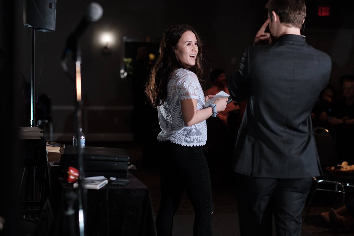 A woman with long hair is smiling and holding a piece of paper, while a man in a suit stands next to her, partially turned away.