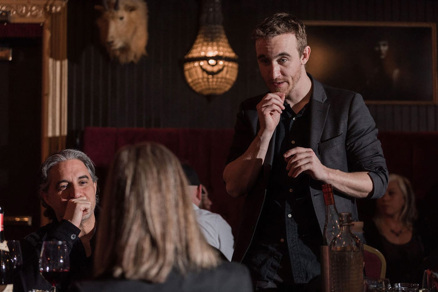 A man in a blazer is speaking to a group at a table in a dimly lit restaurant, while another man with gray hair listens thoughtfully. Wine glasses and bottles are visible on the table.