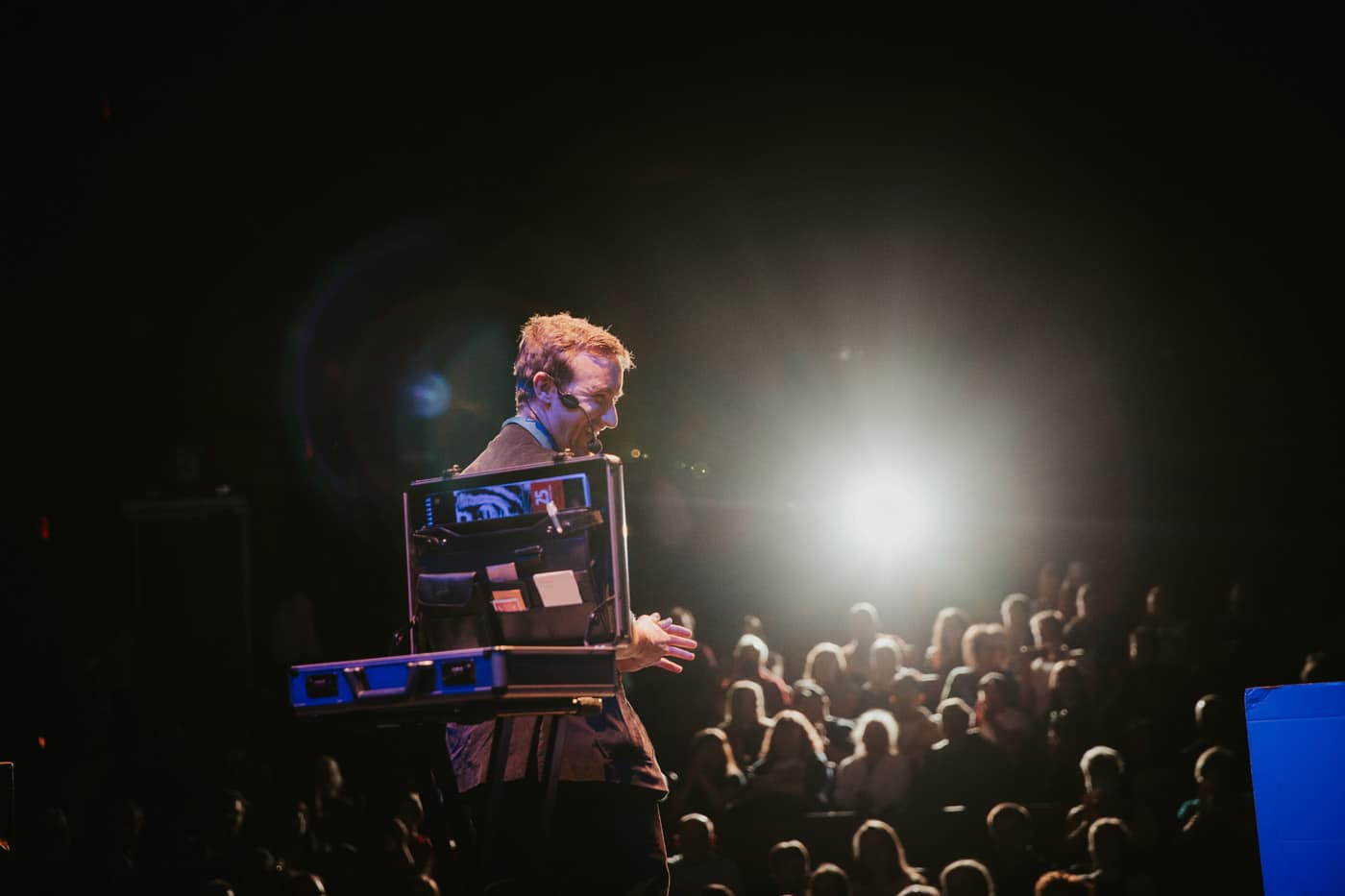 A speaker stands on stage, smiling and engaging with an audience, illuminated by a spotlight. The audience is partially visible in a darkened theater.