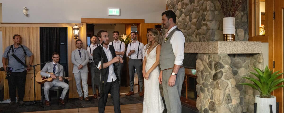 A groom and bride stand together during a wedding ceremony, while a speaker holds a microphone and addresses them. Guests, including musicians, are seated in the background.