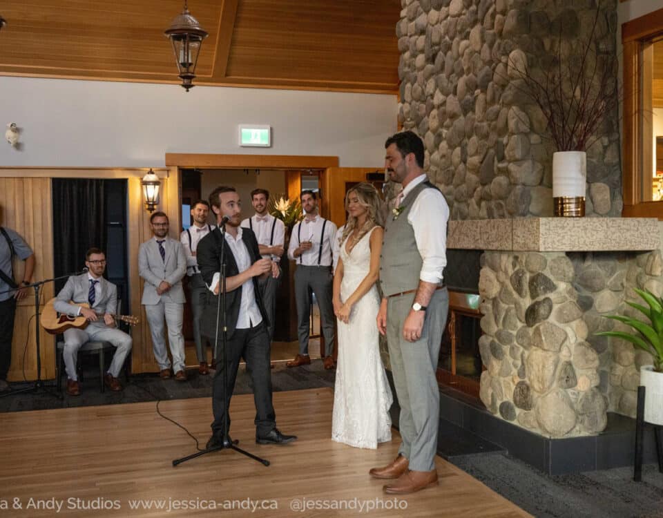 A groom and bride stand together during a wedding ceremony, while a speaker holds a microphone and addresses them. Guests, including musicians, are seated in the background.