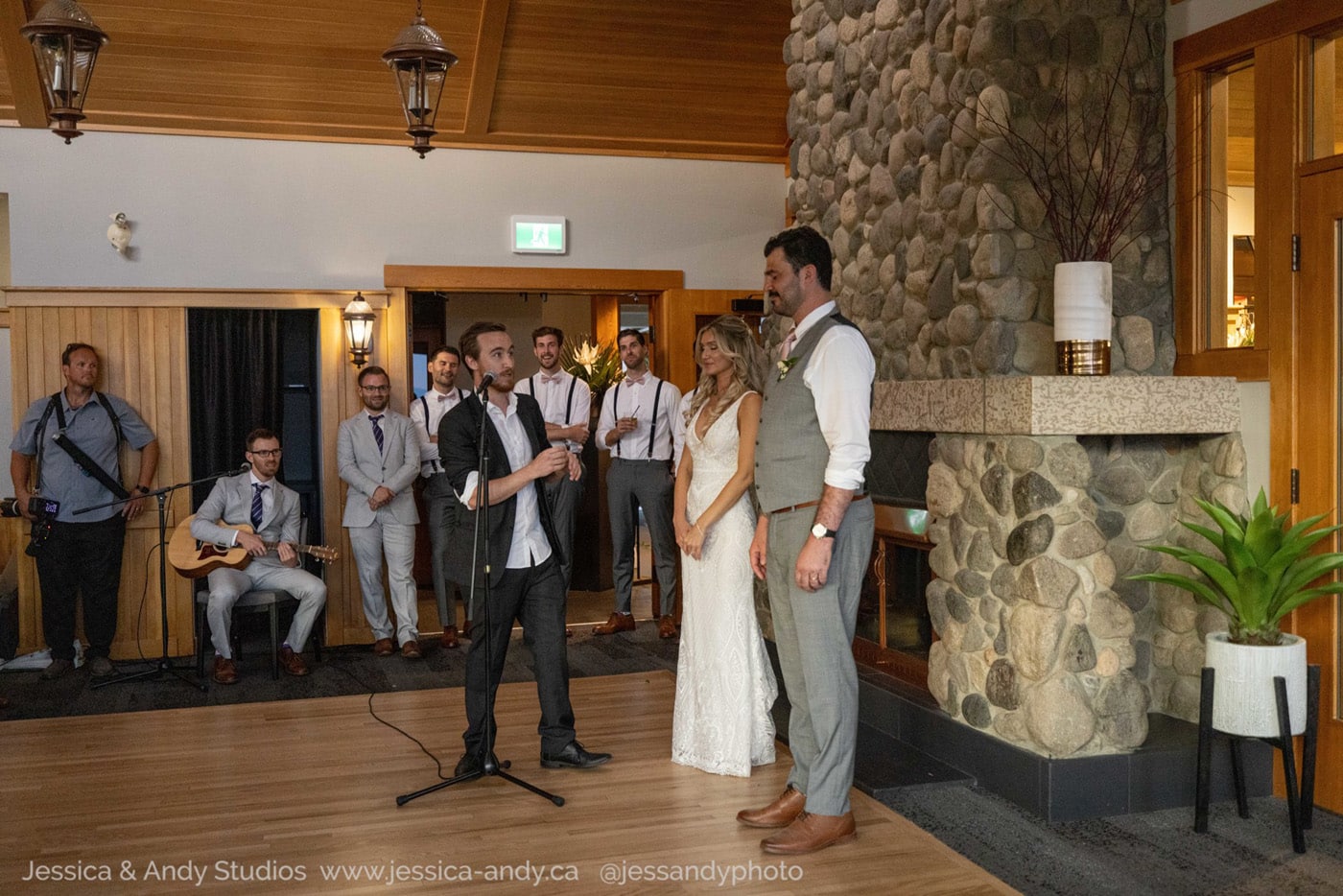 A groom and bride stand together during a wedding ceremony, while a speaker holds a microphone and addresses them. Guests, including musicians, are seated in the background.