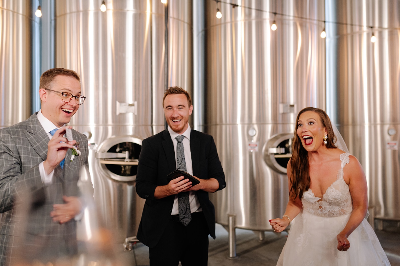 A joyful bride in a wedding dress is celebrating with two men, one in a suit and one in a gray checkered suit, against a backdrop of large silver fermentation tanks.